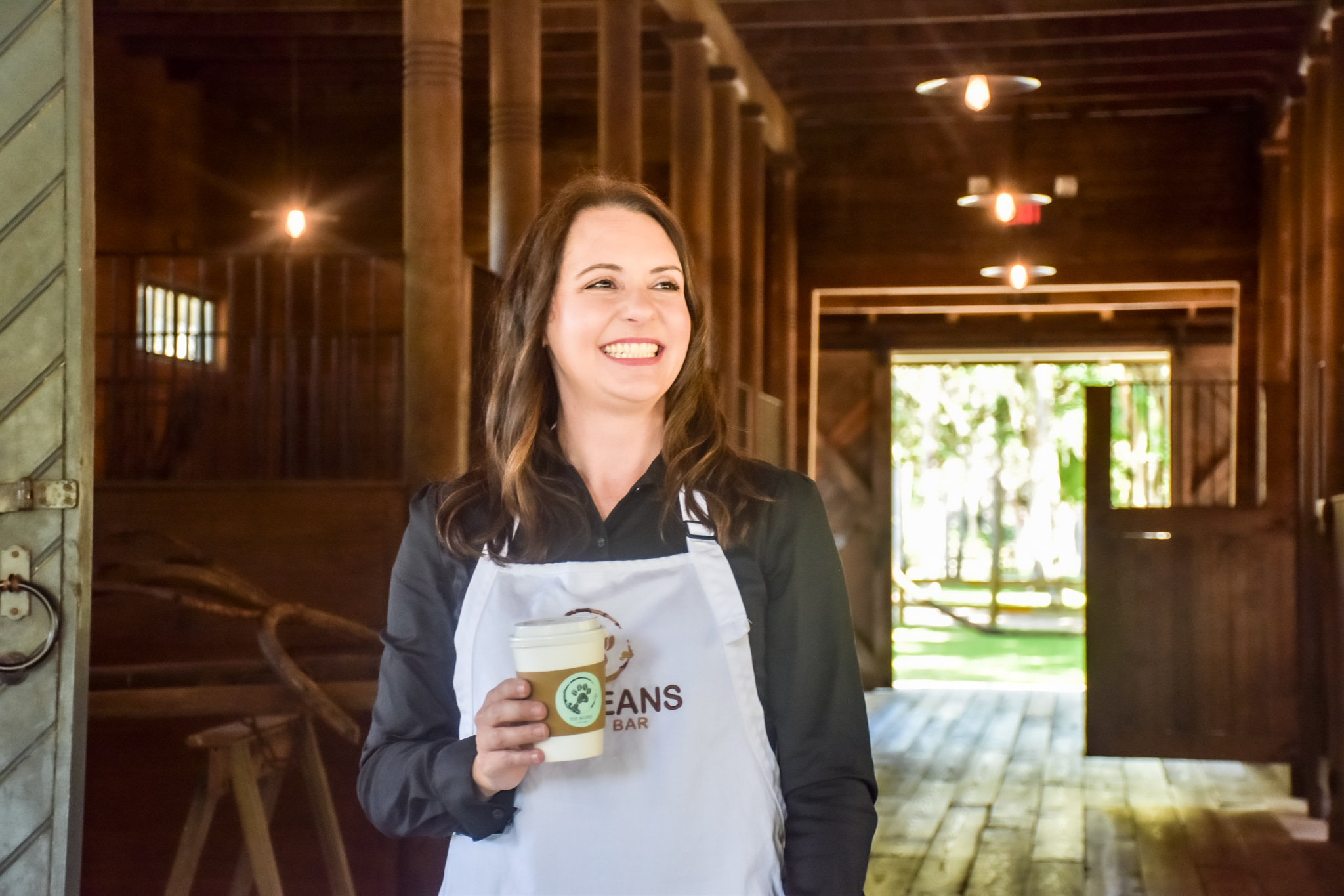 A woman with long brown hair is smiling and holding a disposable coffee cup while wearing a white apron featuring a logo with the text "BEANS." She stands inside a rustic wooden building with warm lighting, as her mobile coffee cart sits just outside, ready for her next coffee catering event.