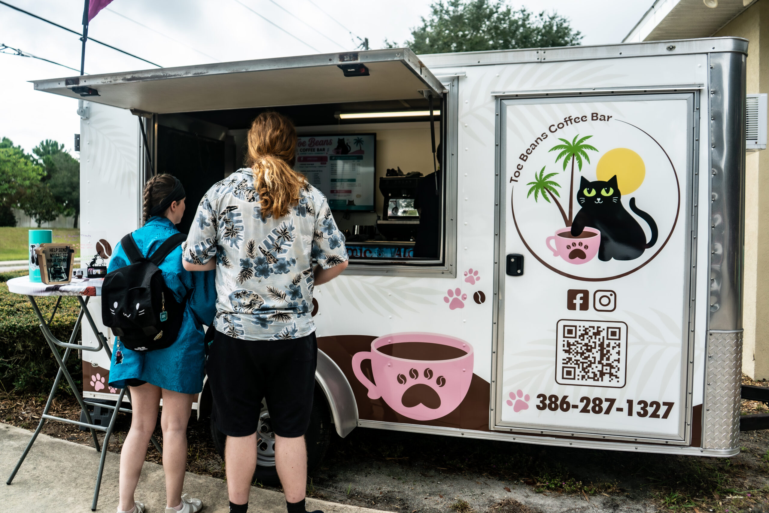A woman with long brown hair is smiling and holding a disposable coffee cup while wearing a white apron featuring a logo with the text "BEANS." She stands inside a rustic wooden building with warm lighting, as her mobile coffee cart sits just outside, ready for her next coffee catering event.